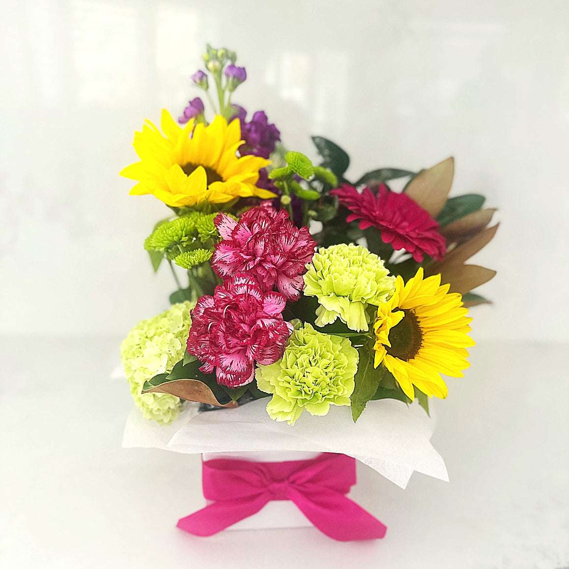 Box arrangement of fresh flowers with sunflowers, carnations, and green foliage tied with a pink ribbon in a white gift box.