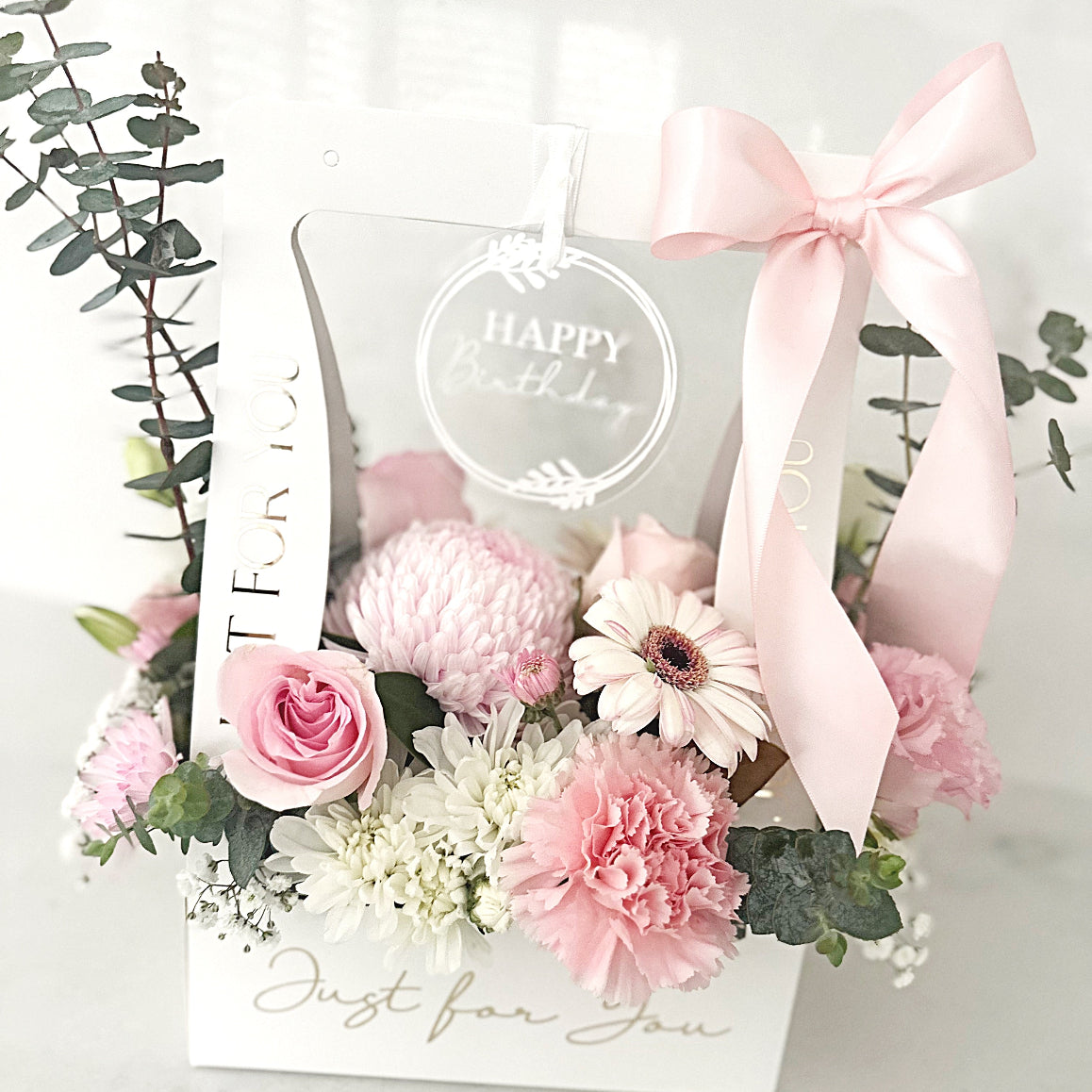 Floral arrangement with pink blooms of roses, chrysanthemums and gerberas with a pink ribbon in a white carry bag arrangement box.