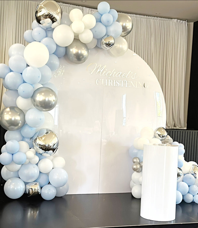 Balloon arch with blue, white, and silver balloons in front of a white curtain.