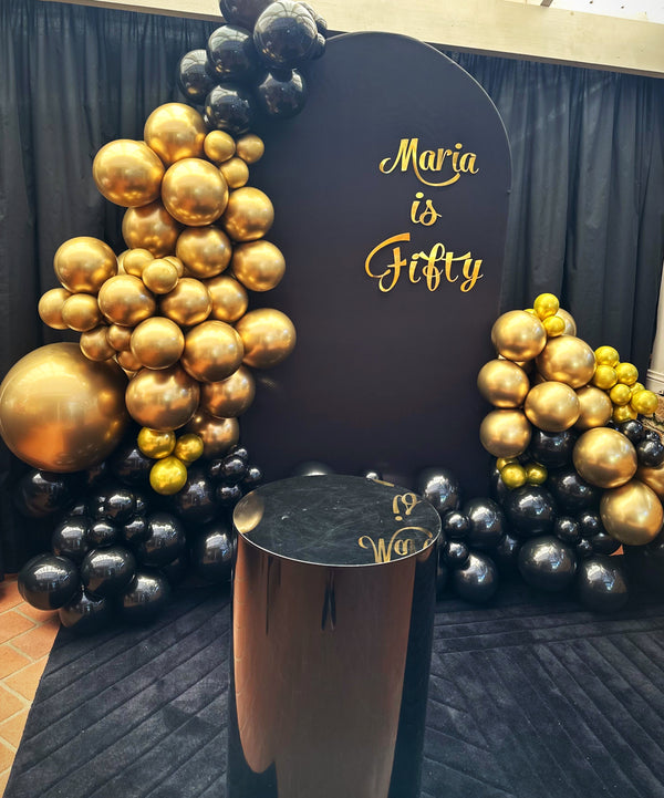 Event setup with gold and black balloons and a sign reading 'Maria is Fifty' on a black arch backdrop and black pedestal.