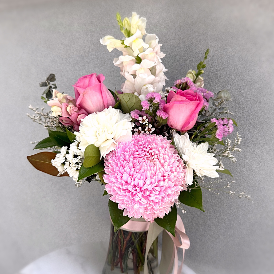 Elegant pink and white flower arrangement features roses and chrysanthemums and snapdragons with luxe green foliage's in a glass vase with pink and sage  ribbons