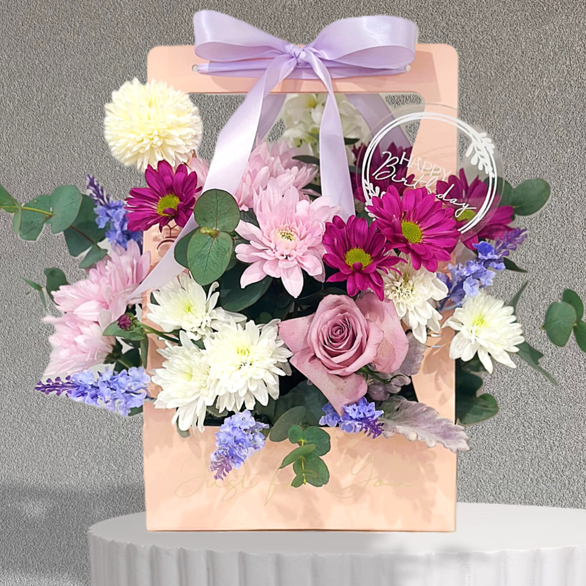 Floral arrangement in a pink Carry Bag Box box with a purple ribbon on a gray background and a Happy Birthday Acrylic Tag