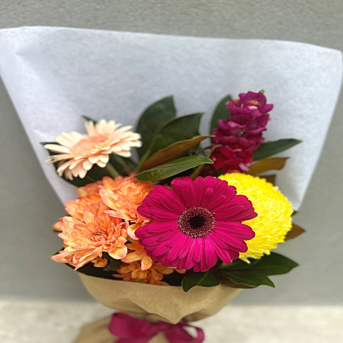 Bouquet of colourful flowers wrapped in brown paper with a pink bow on a light gray background.