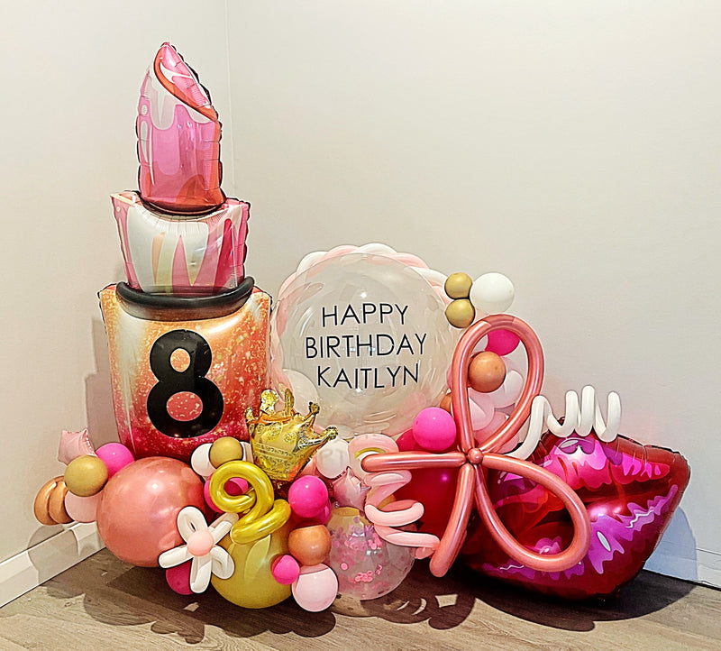 Pink birthday balloon arrangement with a foil lipstick balloon and lips balloon with 'Happy Birthday' on a clear bobo balloon and a white background