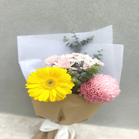 Beautiful posy featuring a yellow gerbera and pink chrysanthemums wrapped in brown paper with a white bow against a gray background